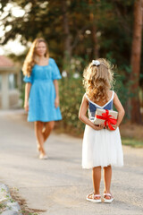 Child daughter hides behind present for mom and Happy Woman in blue dress against green blurred background. Spring family holiday concept. Women's day, Mothers day,Birthday for mom