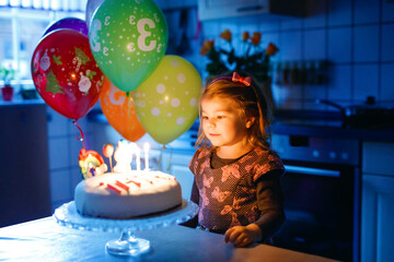 Adorable little toddler girl celebrating third birthday. Baby toddler child with homemade unicorn cake, indoor. Happy healthy toddler is suprised about firework sparkler and blowing candles on cake