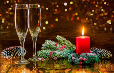 Two glasses of champagne and a burning candle in decorative Christmas wreath of spruce branches on dark background.