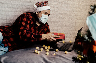 Christmas presents giving. Man in medical mask sharing box gifts through video calls. Holidays in isolation. New year tree and lights with decorations. Family and friends celebrating online.