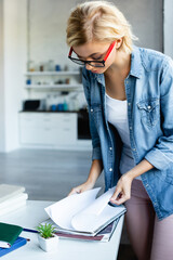 young blonde woman in eyeglasses working from home and turning pages of notebook