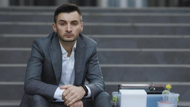 Sad businessman sitting on stairs outdoor with box of stuff as lost business. Fired man. Male office worker in despair lost job. Unemployment rate growing due pandemic