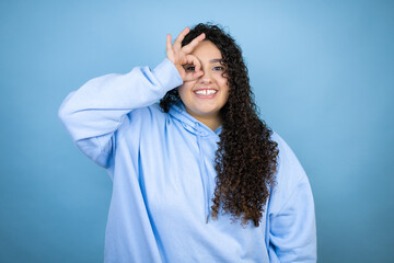 Young beautiful woman wearing casual sweatshirt over isolated blue background doing ok gesture shocked with smiling face, eye looking through fingers