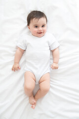 funny healthy baby girl 6 months in white bodysuit lying on bed on white bedding. top view.