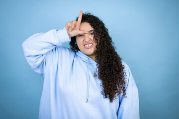 Young beautiful woman wearing casual sweatshirt over isolated blue background making fun of people with fingers on forehead doing loser gesture mocking and insulting.
