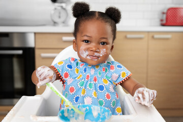Baby girl with messy face eating yogurt with spoon in high chair