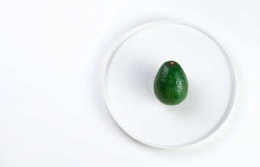 Green unpeeled avocado in a flat white plate on a light background. The view from the top. The concept of wholesome food.