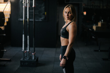 Fototapeta na wymiar Portrait of muscular young woman with perfect athletic body posing during sport workout training at modern gym with dark interior, looking at camera.
