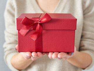 Female hands hold a red gift box. Birthday and Christmas gift with a maroon bow and ribbon. Giving a gift in a box. Time for gifts and Christmas.
