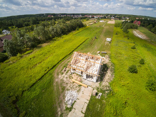 Aerial view on detached house under construction. House in basic state. Located on green plot in small village. Wooden roof structure. Meadows around.
