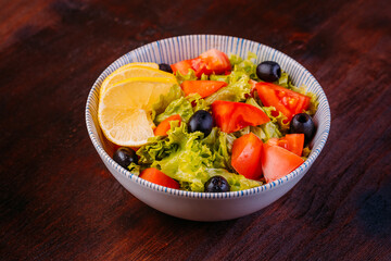 Healthy salad with tomatoes, lettuce, olives, lemon and olive oil in bowl on wooden background. Healthy food, clean eating, dieting, top view.
