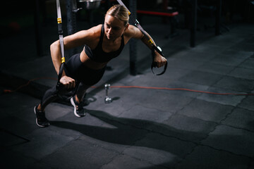 Sporty young woman with perfect muscular body wearing black sportswear working out on simulator during sport training at modern fitness gym with dark interior.