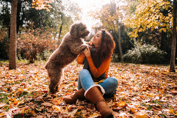 A brunette woman playing with her dog in the park in a sunny autumnal day. The dog has its paws on its owner. She is looking at her dog with love. Lifestyle and outdoors pets in autumn.