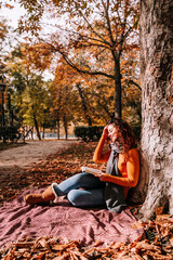 A beautiful woman reading a book in the park. She is sitting and leaning against a tree in a sunny autumnal day. Lifestyle autumnal outdoors.