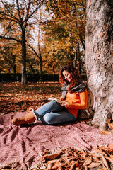 A beautiful woman sitting and leaning against a tree while reading a book in a park in a sunny autumnal day. Lifestyle autumnal outdoors.