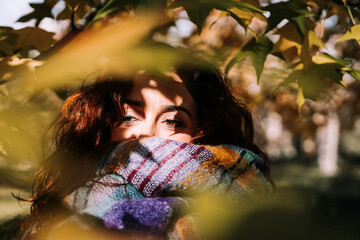 A beautiful brunette woman behind the leaves looking at something among them. She has a nice brown eyes. She is wearing a big scarf covering half of her face. Autumnal portrait close up.