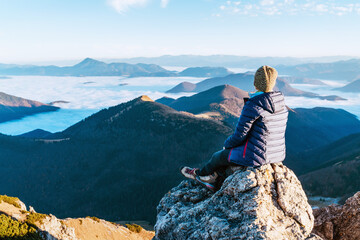 Young hiker woman sitting on the mountain summit cliff and enjoying mountains valley covered with clouds view. Successful summit climbing concept image.
