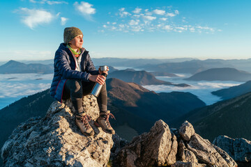 Young hiker woman sitting on the mountain summit cliff, drinking tea from a thermos flask and...