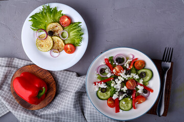 White plate with grilled zucchini with cherry tomatoes on lettuce and Greek salad on a napkin with a fork near vegetables.