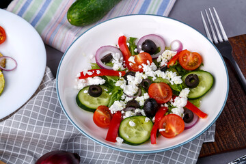 White plate with grilled zucchini on lettuce leaves and Greek salad on a gray table on a napkin with a fork near vegetables.