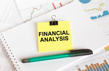 A yellow sticker with text Financial Analysis is in a Notepad with a green pen financial charts and documents