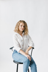 Beautiful blonde girl in white shirt and jeans sitting on a chair on a white background in the studio.