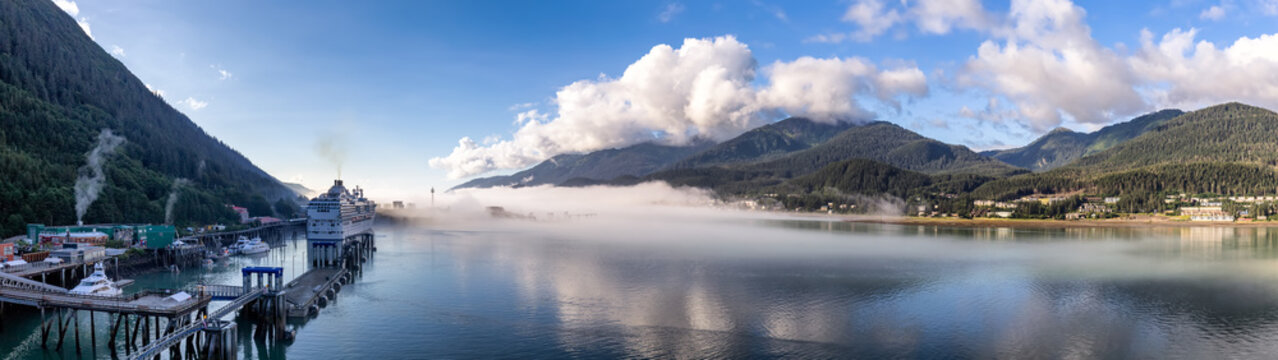 Panoramic shot of port of Juneau and mountains covered with clouds and fog in Gastineau Channel, Alaska. Cruise ship and boats docked in the port. Blue cloudy sky as a background