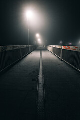 Moody and misty path (sidewalk) in the dark city at night with white street lamps. Road with lane and railling - depth field vertical photo. Empty foggy bridge with graffiti on the construction.