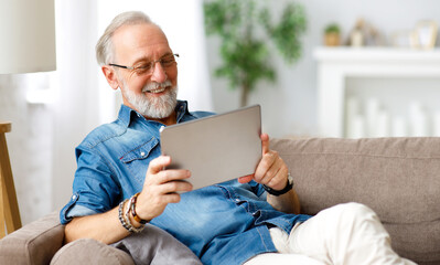 Cheerful aged man using tablet on couch.