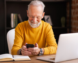 Cheerful aged freelancer uses a smartphone in home office.