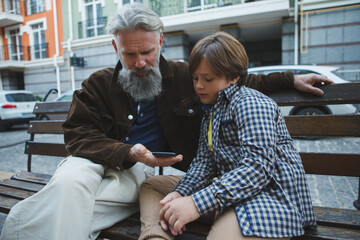 Elderly man using smart phone with the help of his young grandson, sitting on a bench in the city