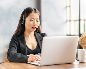Happy asian businesswoman in headset speaking by conference call and video chat on laptop in office, chinese support service agent consulting customer