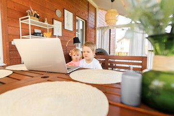 Small smiling kids brother and sister laugh and play laptop, communicate video conference chatting. Spacious cozy home interior.