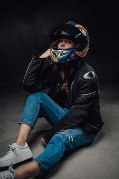 Wearing helmet female moto racer dressed in black jacket with ripped jeans poses in dark background sitting on floor.