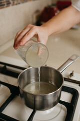 A close photo of a hand of a woman who is pouring fresh lemon juice from the glass to the saucepan on the gas stove