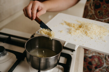 A close photo of a hand of a woman who is throwing lemon zest off the board with a knife to fresh lemon juice in the saucepan on the gas stove