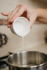 A close photo of a hand of a young woman who is pouring cream from the cup to the saucepan with lemon juice and zest on the gas stove