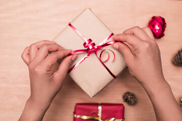 Top view photography of woman's hands tying red ribbon on handmade present on wood table, package for christmas gifts.