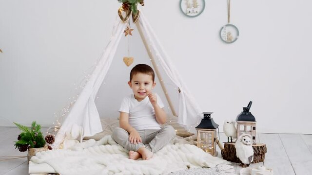 Little caucasian boy laughing while lying in a toy kids teepee in a white scandinavian christmas interior.	