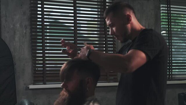 Blurry image of a Barber who makes a fashionable haircut for a male client. In the background is a large window with blinds.