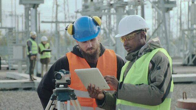Bearded power engineer and African-American worker in helmets check data by level-theodolite at blurred electric substation closeup