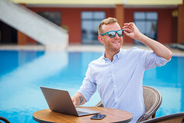 Young male businessman in sunglasses works at a laptop sitting at a table near the pool. Remote work. Freelancer