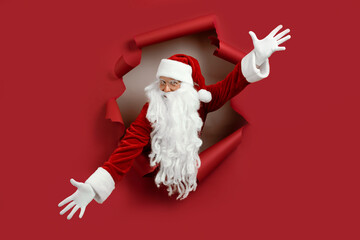 Santa man spread his arms in different directions and look emotionally through a paper hole. Bearded man in santa hat looking through hole on red paper.