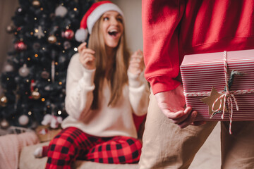 Surprising her... Close-up rear view of man in red sweater holding a Christmas gift box behind his back while standing near decorated christmas tree.