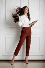 A beautiful young girl reading a book and throwing long wavy hair up. Model in a white blouse and red trousers in office style clothes.
