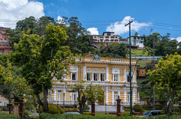 Petropolis, Brazil - December 23, 2008: Palacio Rio Negri, or black river Palace frontal view with busy street in front and surrounded by green trees. Houses higher up on hill under blue cloudscape.
