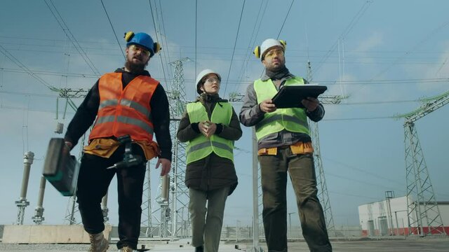 Construction engineers and woman brigadier in helmets walk along electric power substation and discuss project low angle shot