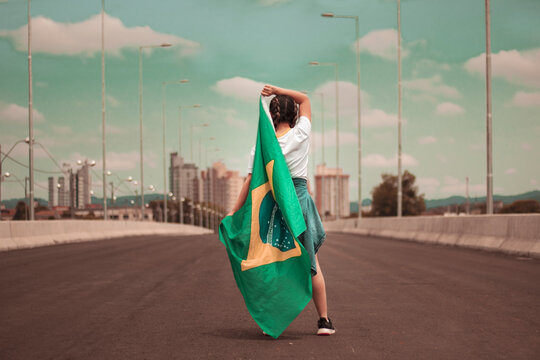 Mulher em cima de uma ponte, com a cidade ao fundo, trajando blusa branca e camisa na cintura, segurando a bandeira do Brasil e sorrindo