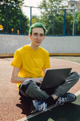 Fototapeta premium Freelancer, it professional in free space, remote working at online platform for programmers. Smiling young male freelancer with green hairstyle working on laptop in outdoor street.