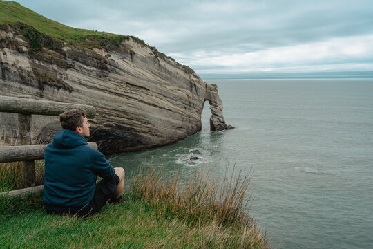 Young caucasian man sitting on top of the cliff and looking at Cape Farewell rock formation. New Zealand